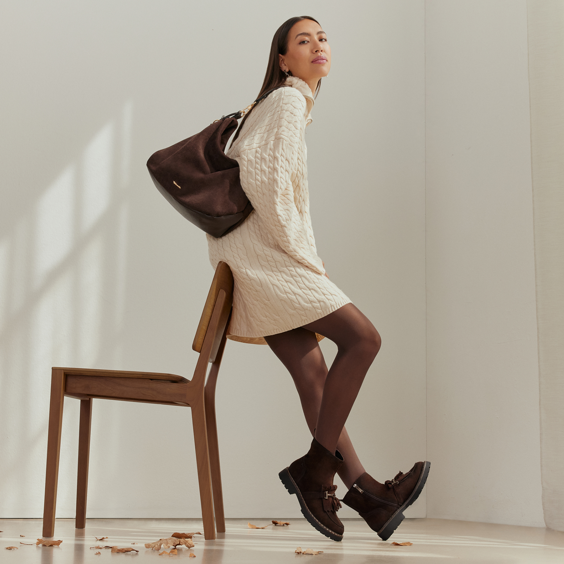 Woman in a cream knitted dress, brown ankle boots and dark tights leaning against a wooden chair, wearing a brown shoulder bag.