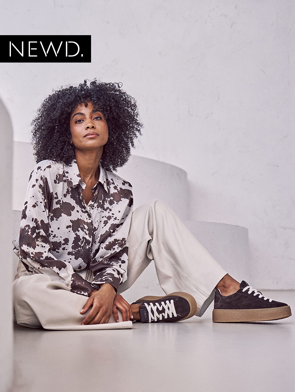 Modell in gemusterter Bluse und heller Hose sitzt auf dem Boden, tr&auml;gt zweifarbige Sneaker. Nat&uuml;rliche Lockenfrisur und minimales Make-up.