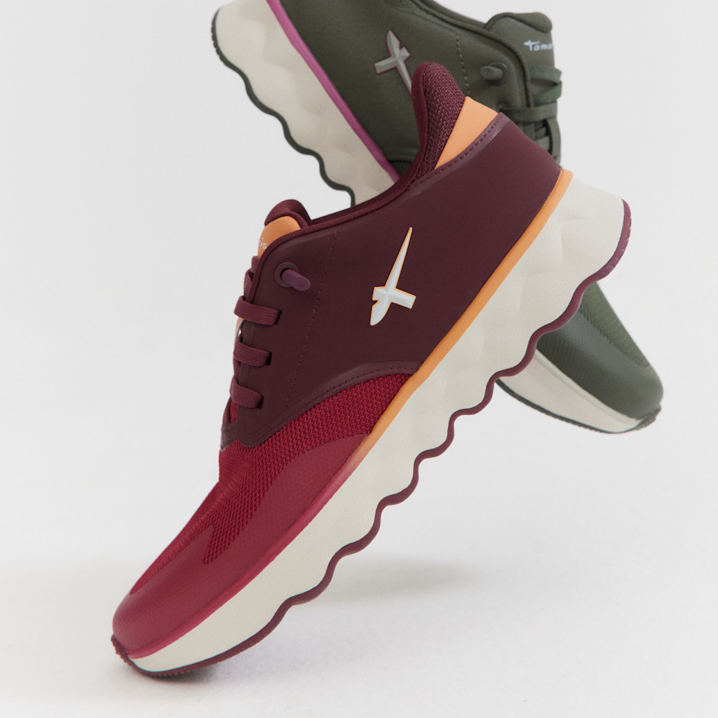 Sporty sneakers in wine red and olive green with a white wavy sole and orange accents, photographed in a dynamic perspective.