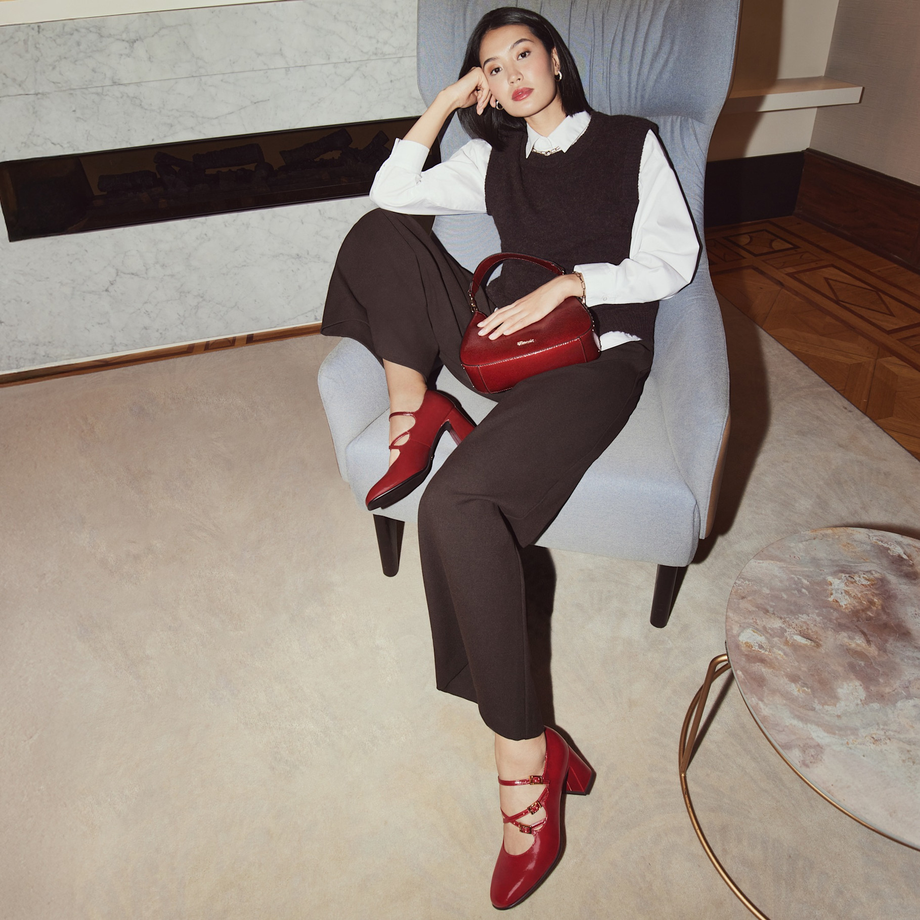 Donna indossa un elegante completo da lavoro color marrone scuro in combinazione con scarpe rosse e borsa rossa. Seduta su una poltrona grigia in una stanza minimalista.