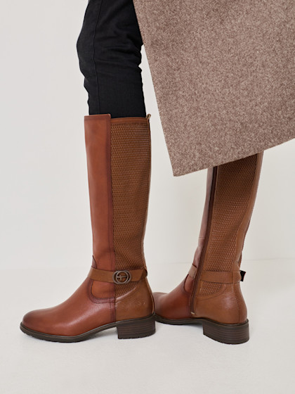 Elegant brown leather riding boots with buckle and perforated shaft, worn with black trousers and beige coat