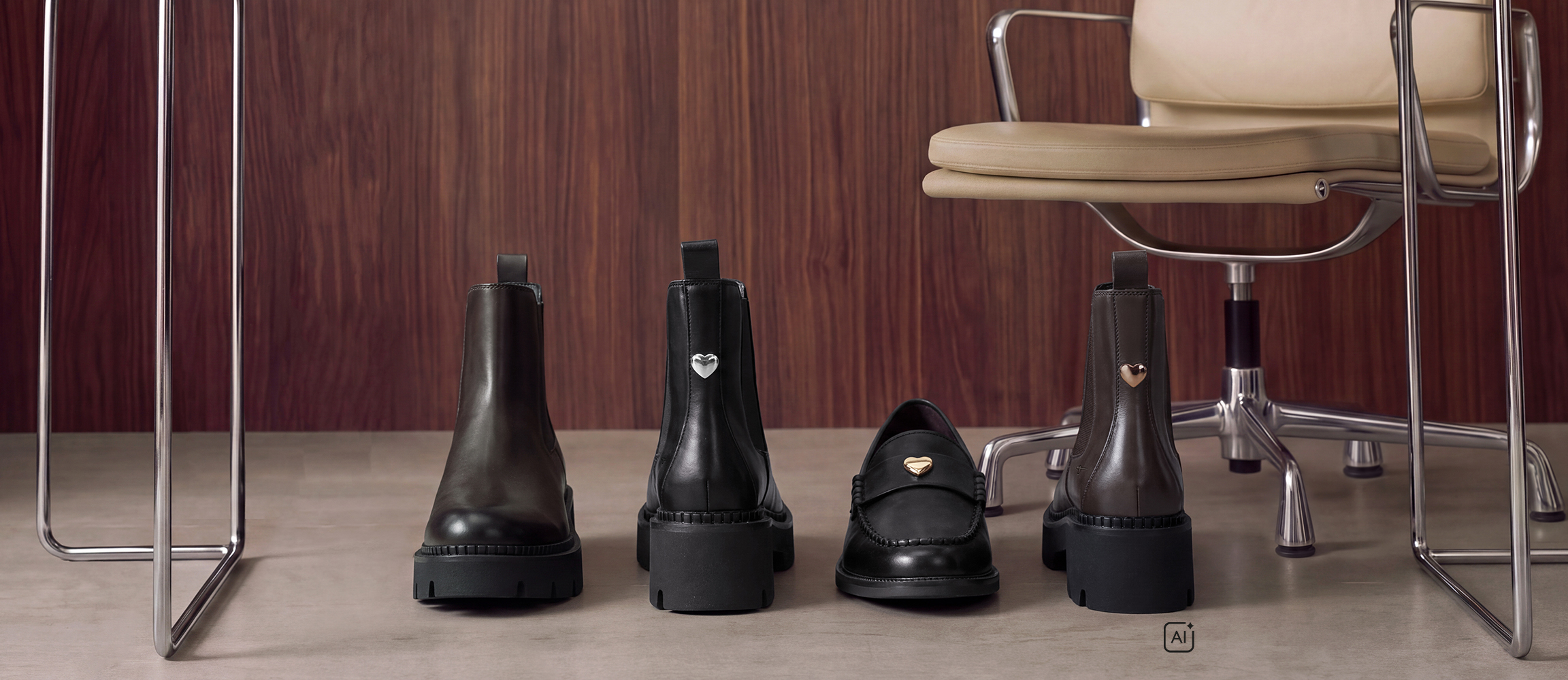 Drie zwarte leren schoenen: twee Chelsea‑boots met hartdetail en &eacute;&eacute;n loafer, op een grijze vloer voor een houten wand met een beige stoel.