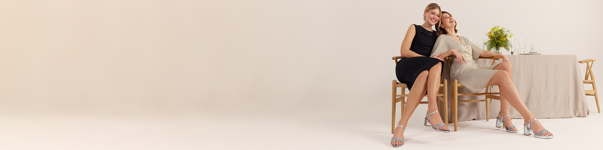 Twee vrouwen in elegante jurken zitten ontspannen op houten stoelen naast een tafel gedekt met gele bloemen