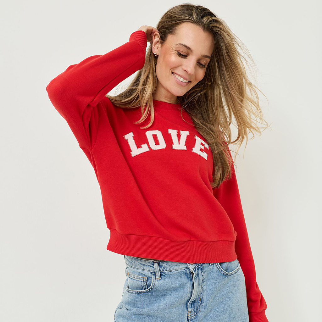 Smilende kvinde i r&oslash;d LOVE-sweater og jeans foran hvid baggrund, med flagrende blondt h&aring;r.