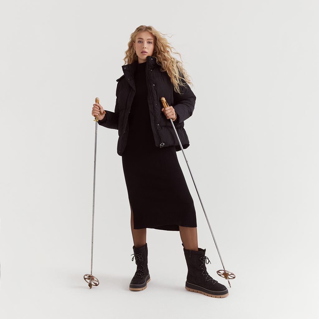 Model in zwarte winterkleding met donsjack, gebreide jurk en laarzen poseert met skistokken voor een witte achtergrond.