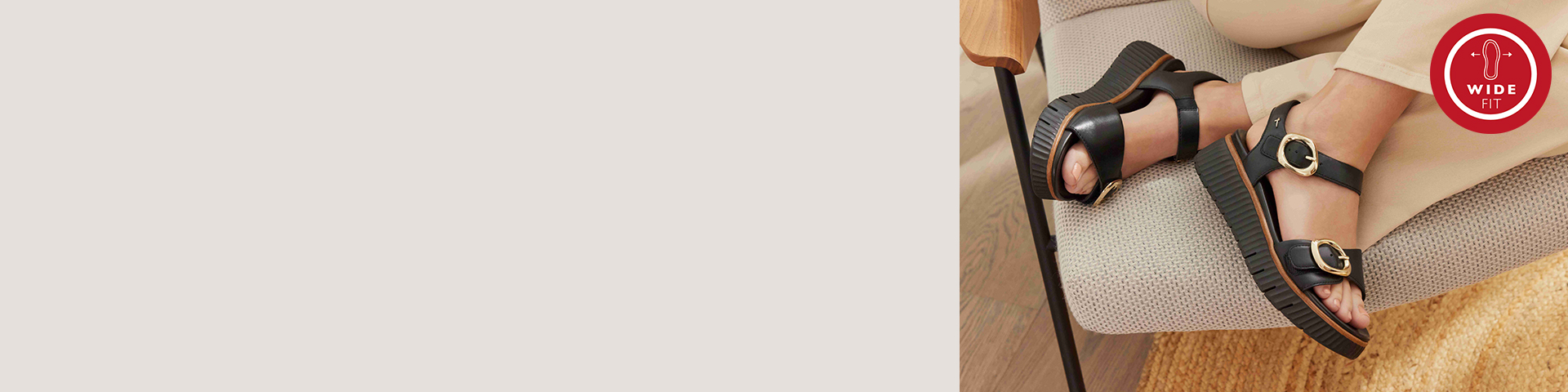 Černé sandály se zlatými přezkami na béžovém čalouněném nábytku, s logem Wide-Fit v rohu.