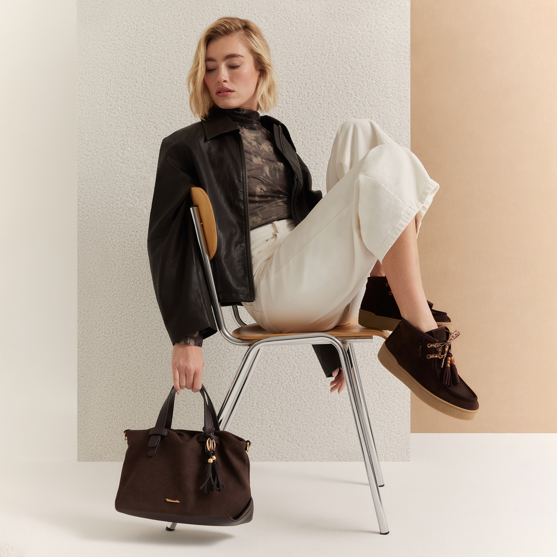 Woman sitting on a chair wearing a black leather jacket, white trousers and brown sneakers, holding a dark brown handbag.