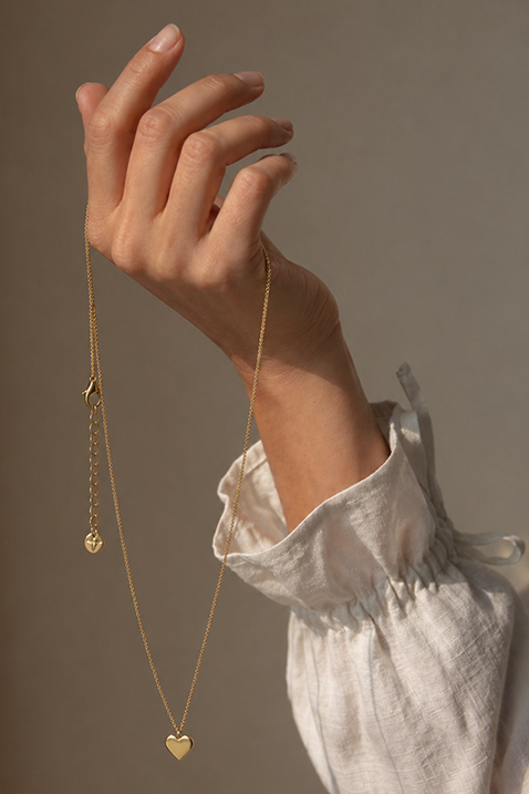 Een hand houdt een fijne gouden ketting met een hartvormige hanger vast. De persoon draagt een witte blouse.
