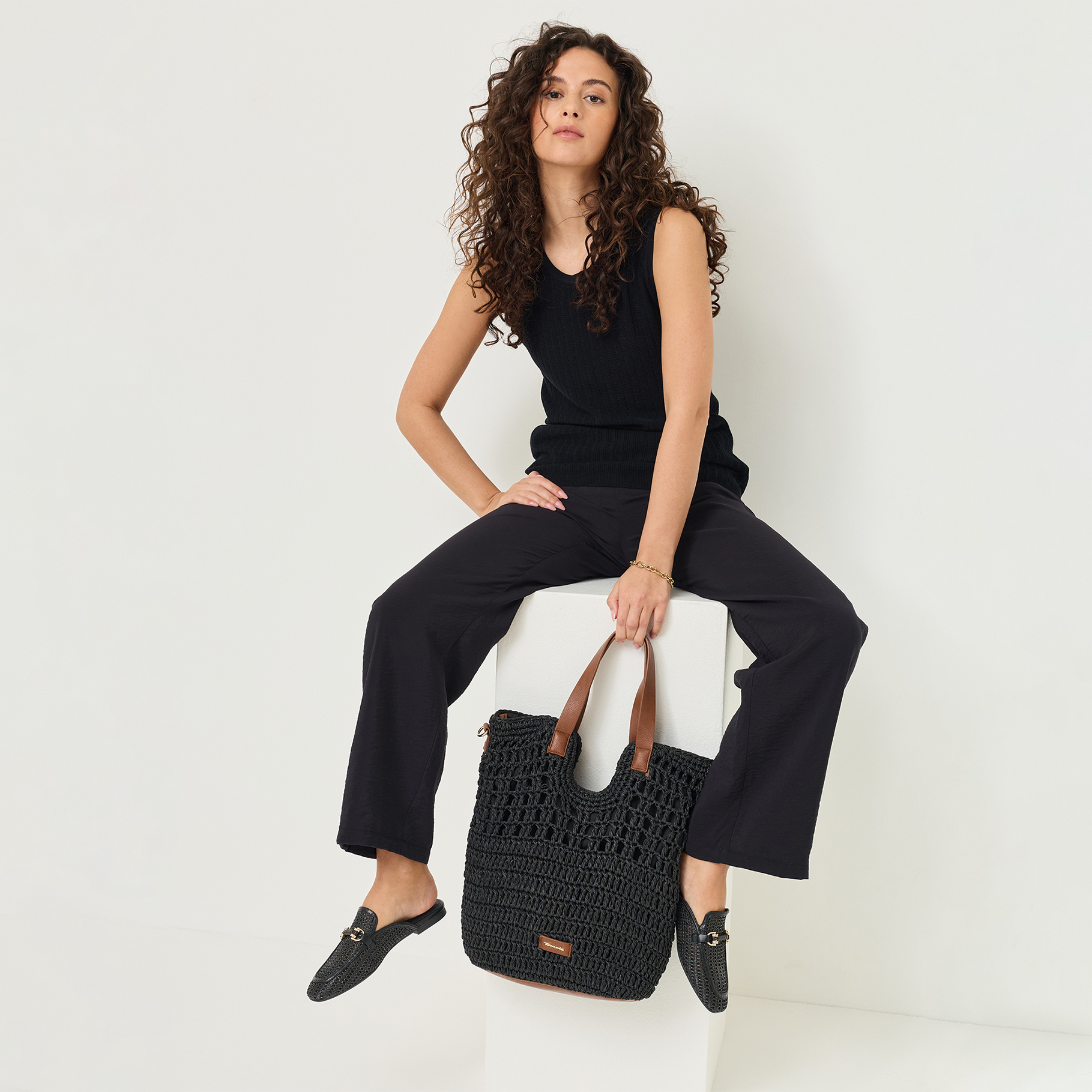 Frau in schwarzem Outfit sitzt auf einem Hocker und h&auml;lt eine schwarze H&auml;keltasche mit braunen Ledergriffen.