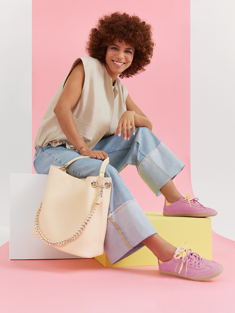 Modisches Outfit mit beiger Bluse, Jeans, rosa Sneakers und cremefarbener Handtasche vor pastellfarbenem Hintergrund.