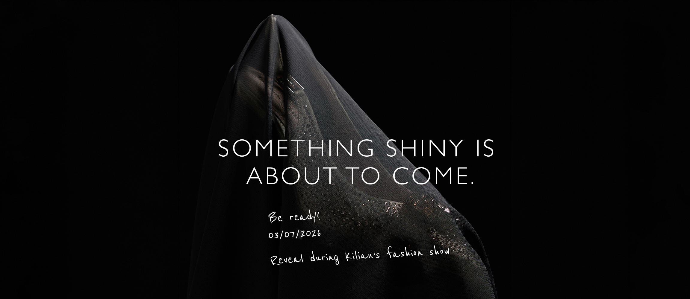 Donkere teaser-afbeelding met een bedekt object. Tekst: Something Shiny Is About To Come. Onthulling tijdens Kilians modeshow op 03-07-2026.