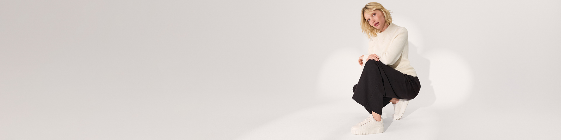 En person i hvid sweater og sorte bukser poserer dynamisk foran en lys baggrund, iført hvide sneakers.