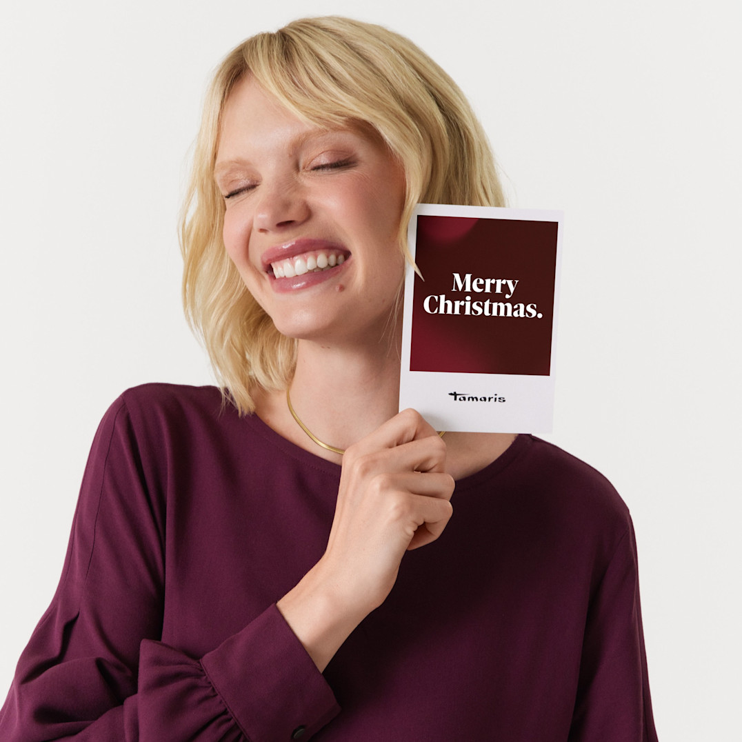 Lächelnde Person in burgunderrotem Kleid hält eine Geschenkkarte von Tamaris mit der Aufschrift "Merry Christmas"