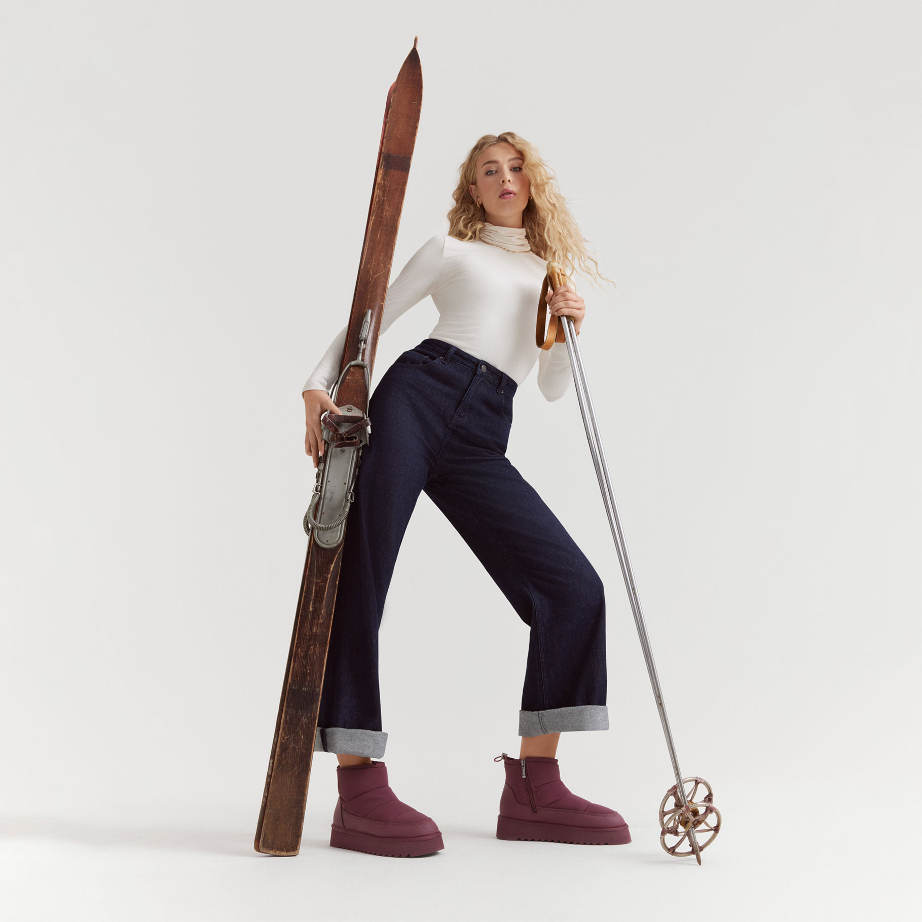 Modelka w białym golfie i ciemnych dżinsach pozuje z nartami vintage, nosi bordowe buty na platformie.