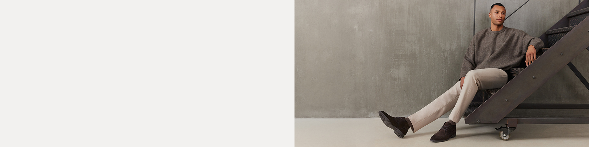 Man sitting casually on a metal staircase, wearing a grey jumper, beige trousers and dark brown boots in front of a grey concrete wall.