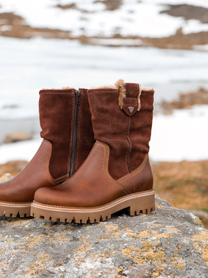 Braune Lederstiefel stehen auf einem Felsen vor verschneitem Hintergrund.