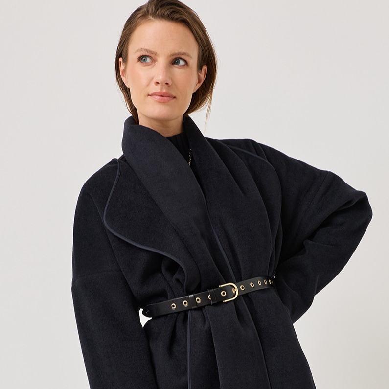 A woman in an elegant black coat with belt poses in front of a white background in a fashionable pose.