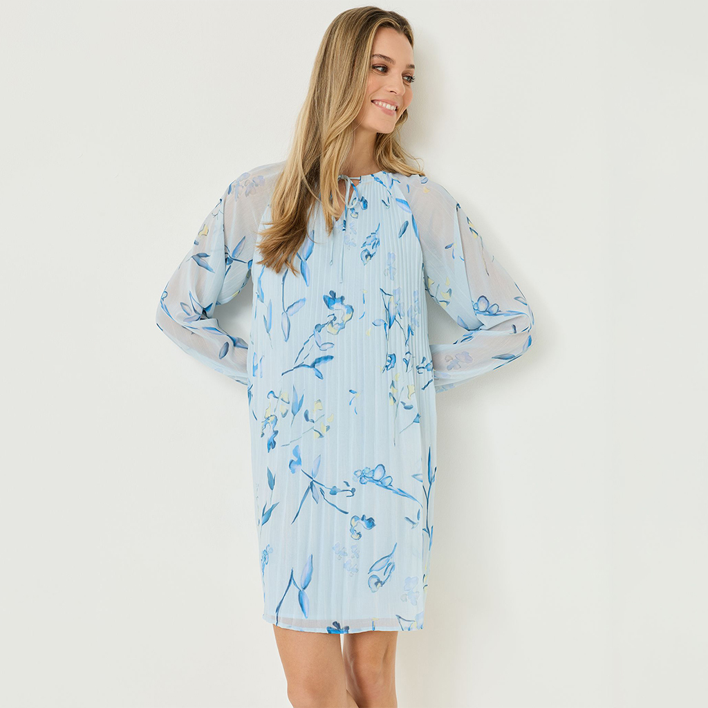 Smiling woman in a light blue pleated dress with floral pattern and sheer sleeves, knee-length design.