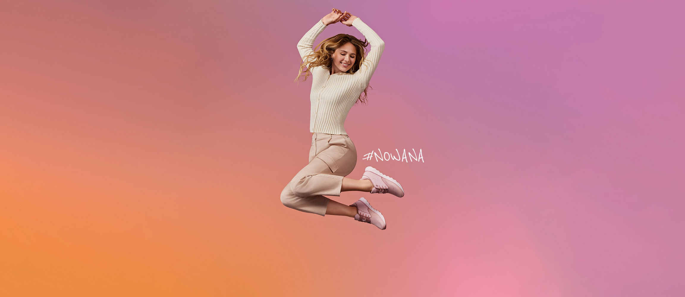 Kvinde i cremefarvet outfit springer glædeligt foran en rosa-orange baggrund, bærer rosa sneakers og #NOWANA skrift er synlig