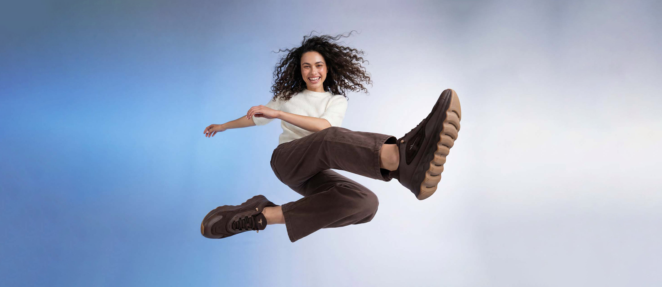 Femme souriante aux cheveux boucl&eacute;s sautant en l'air, portant un pull blanc, un pantalon marron et des baskets marron sur fond bleu.