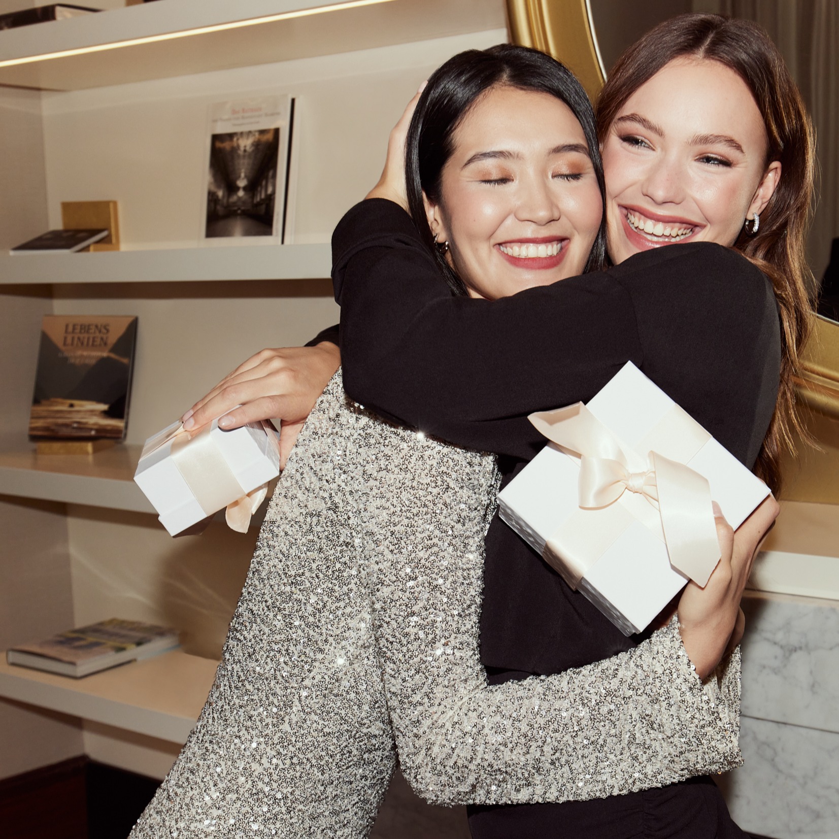 Two friends embrace joyfully, one wearing a black top, the other a glittering sequin dress.