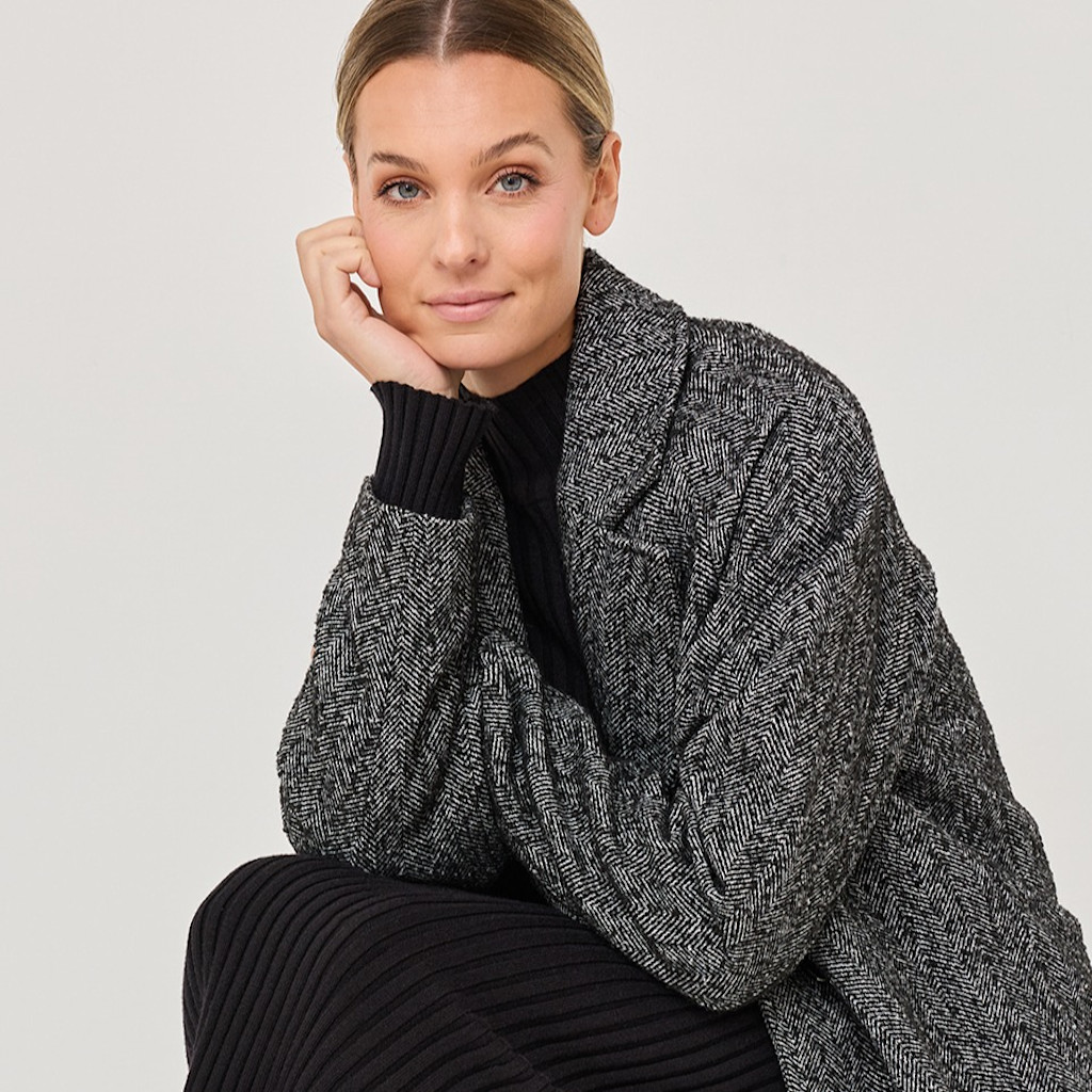 Portr&auml;taufnahme einer Person in grauem Tweed-Mantel und schwarzem Pullover vor wei&szlig;em Hintergrund, die Hand nachdenklich am Kinn.