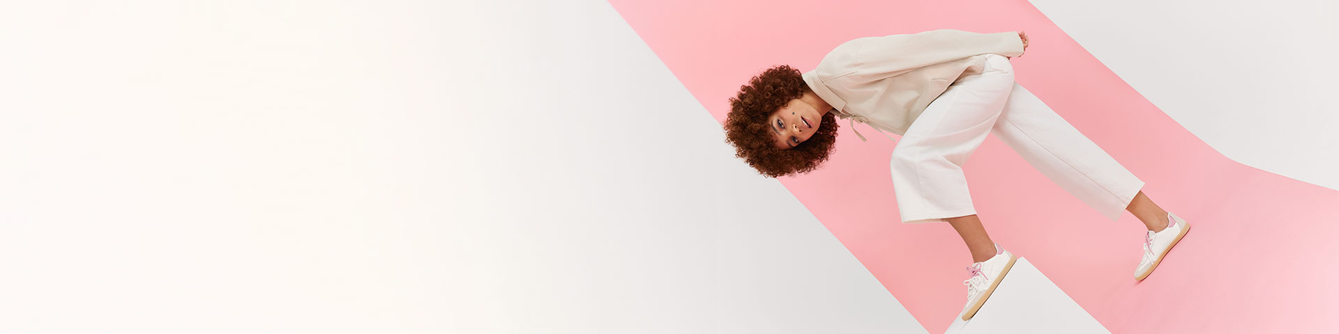 Frau in cremewei&szlig;em Outfit in dynamischer Pose vor rosa Hintergrund, lockiges Haar in Bewegung, wei&szlig;e Sneaker an den F&uuml;&szlig;en.