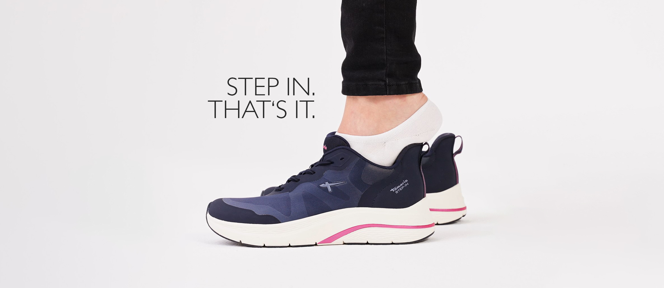 Navy blue sports shoes with a white sole and pink accents, worn with black trousers, with the text "STEP IN. THAT'S IT."