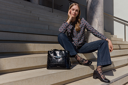 Une femme en chemisier à motifs et en jeans est assise de manière détendue sur un large escalier en pierre devant un bâtiment. Elle porte des escarpins noirs et un sac noir.