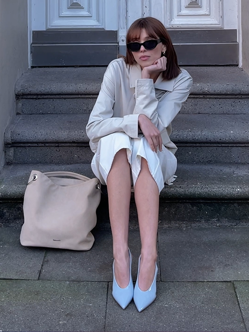 Woman sitting on stairs wearing a beige outfit, light blue pumps and sunglasses, with a beige handbag next to her.
