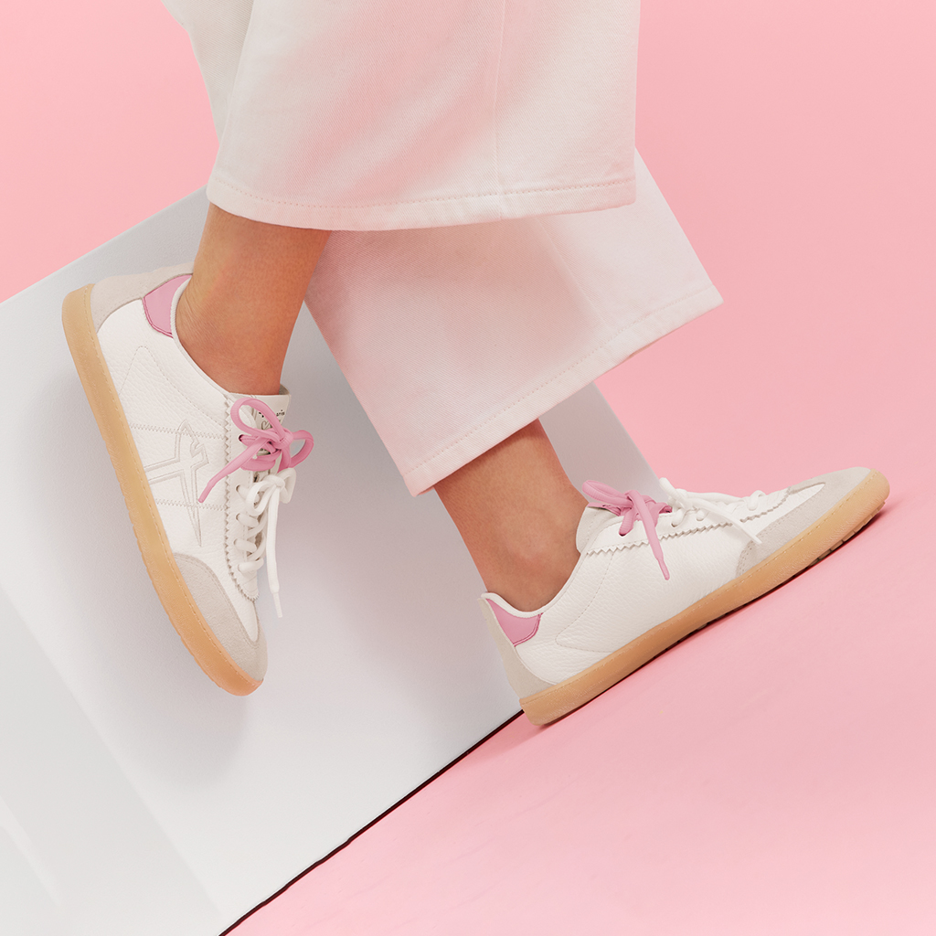 Witte sneakers met roze accenten en rubberen zool, gedragen met een witte broek tegen een roze-witte achtergrond.