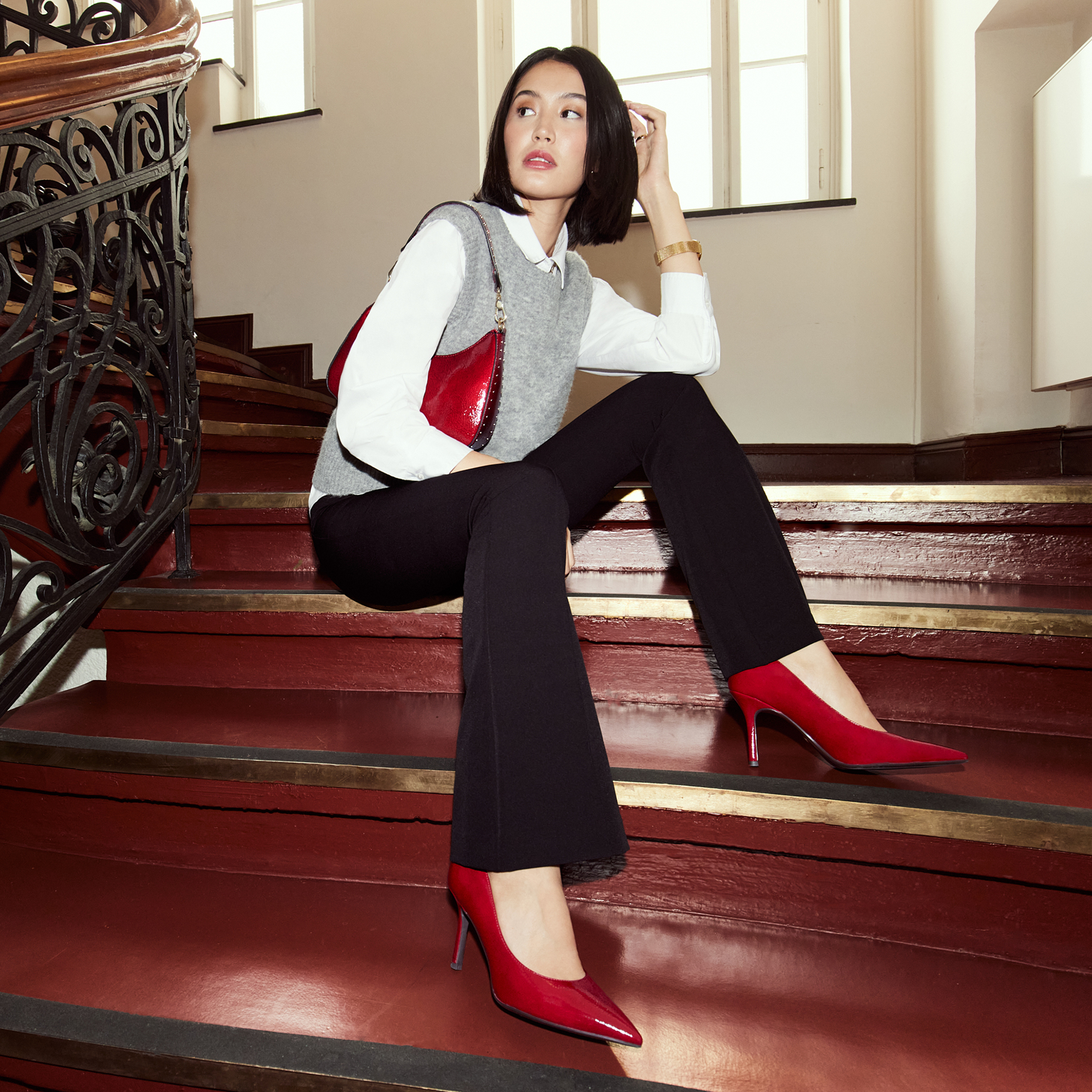 Modell in Business-Outfit mit weißer Bluse, grauer Weste und schwarzer Hose sitzt elegant auf einer Treppe mit roten High Heels.
