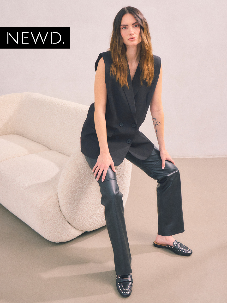 Il modello in gilet nero senza maniche e pantaloni in pelle siede su un divano color crema e indossa mocassini neri impreziositi.