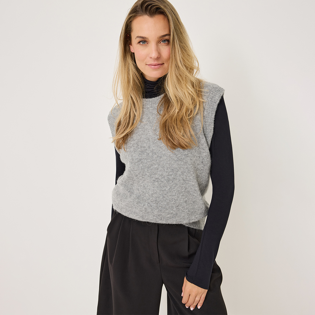 Modello in gilet grigio su maglione nero a collo alto e pantaloni scuri su sfondo bianco