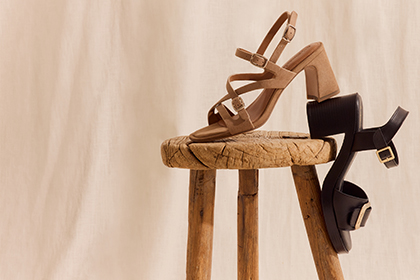 Two elegant women&rsquo;s heeled sandals &ndash; one beige and one black &ndash; on a rustic wooden stool against a light background.
