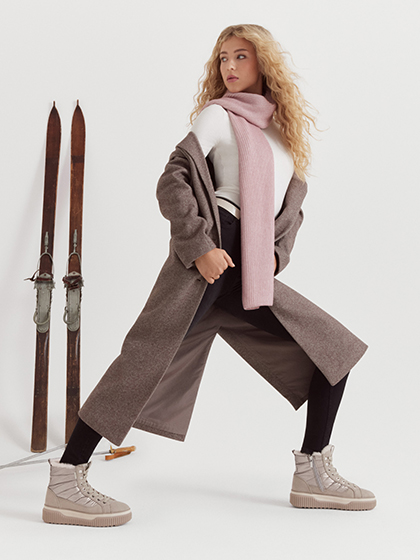 Frau in braunem Wintermantel, rosa Schal und beigen Schneeschuhen posiert dynamisch mit Vintage-Skiern vor wei&szlig;em Hintergrund.