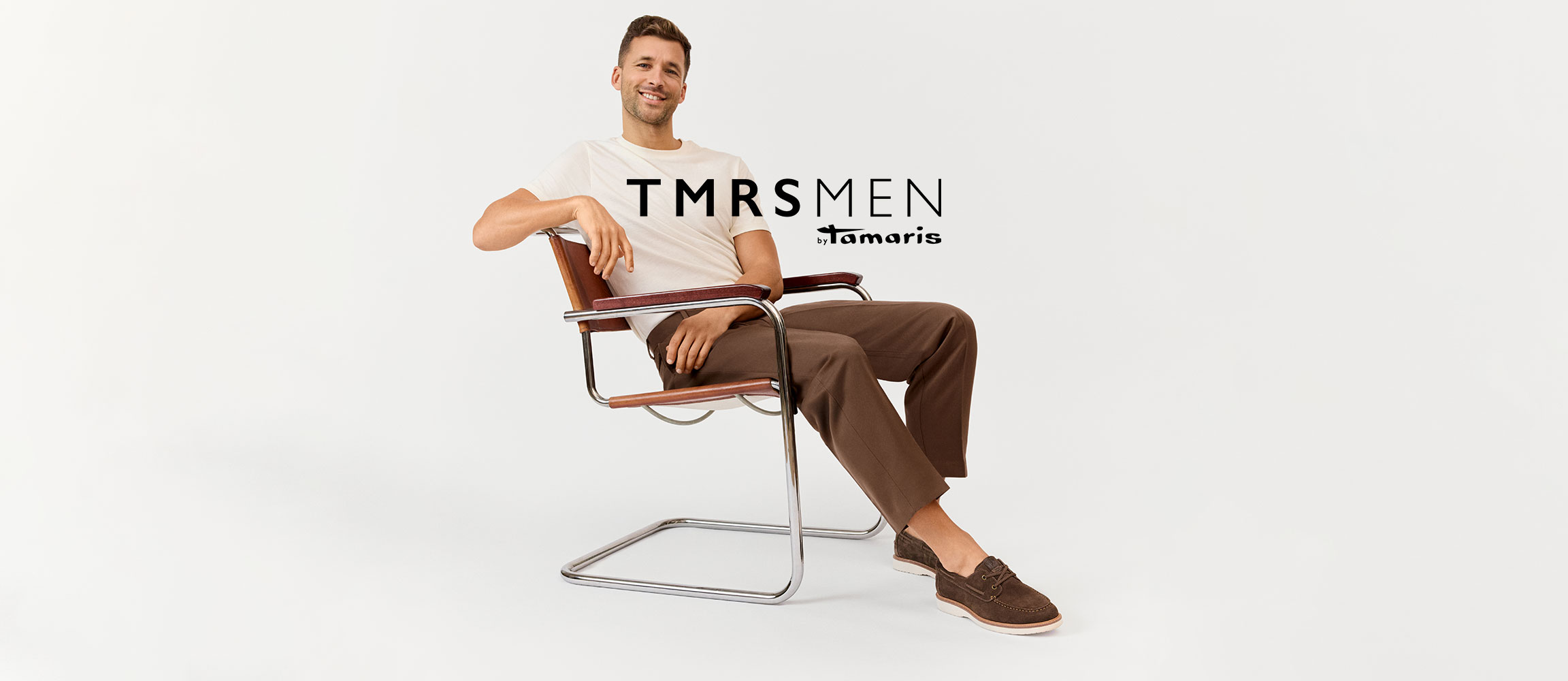 Modell i vit T-shirt och bruna byxor sitter leende i en modern kromstol, TMRSMEN-logotyp i bakgrunden.
