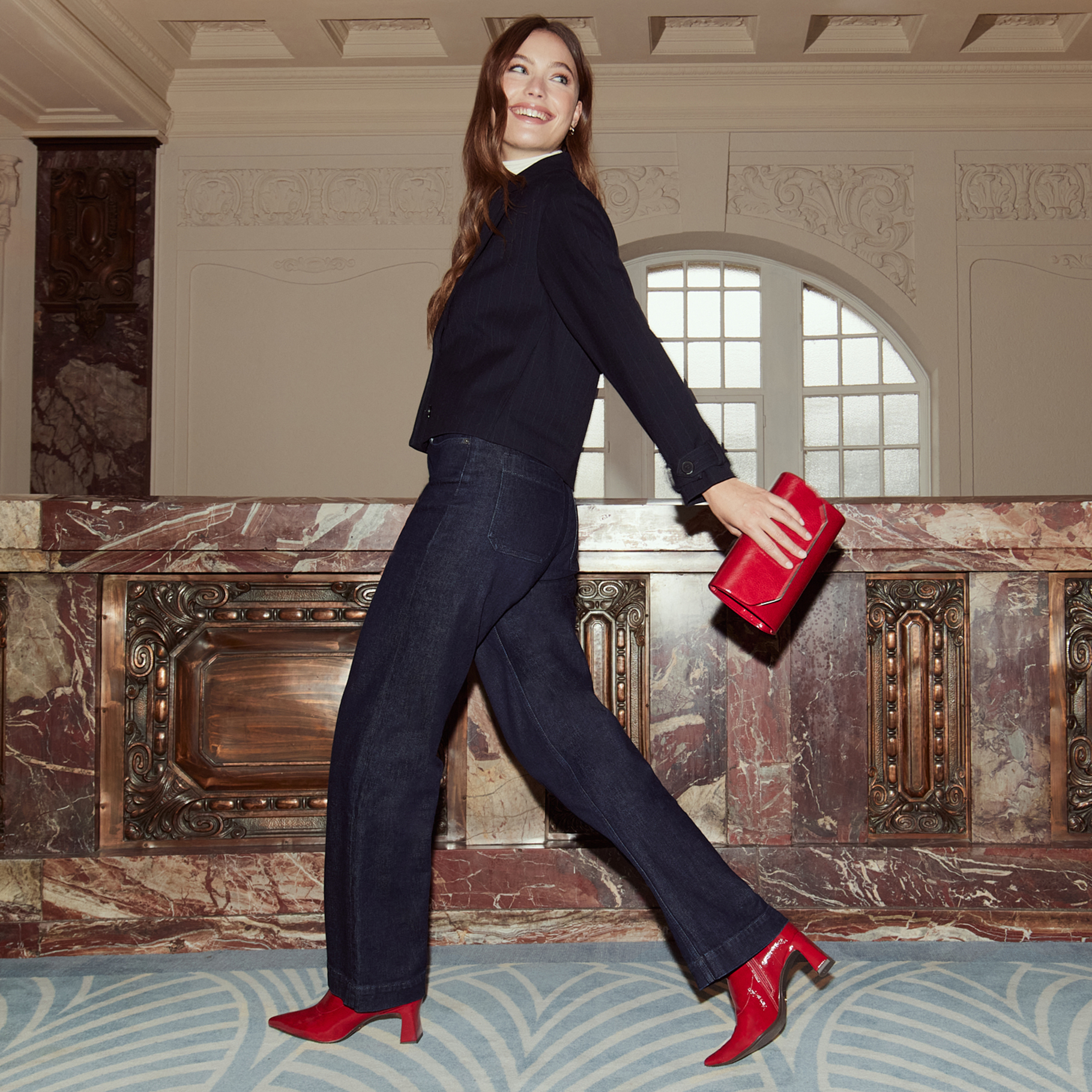 Une femme en tenue sombre avec des chaussures rouges et un sac à main danse de manière dynamique dans une pièce élégante avec des colonnes en marbre.