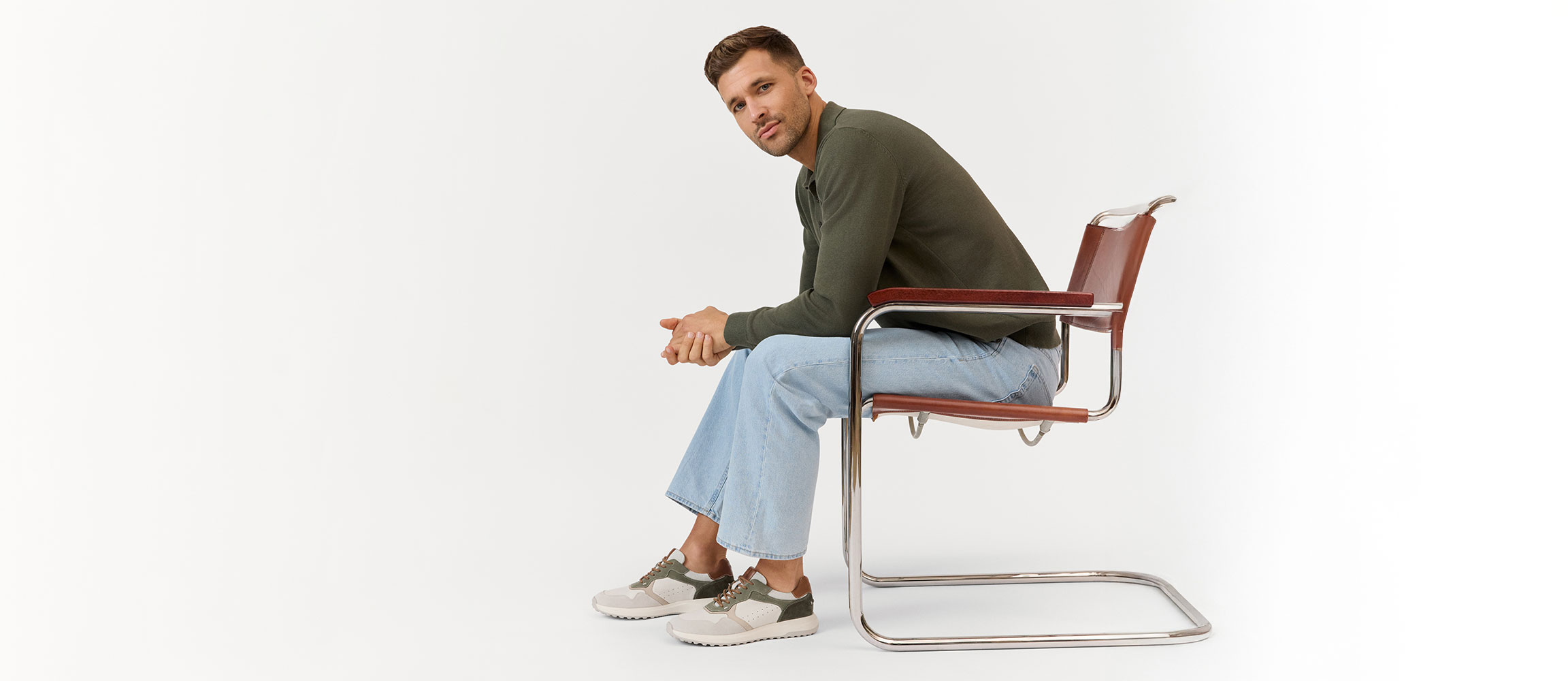 Person in olivgr&uuml;nem Pullover und heller Jeans sitzt seitlich auf einem modernen Stuhl vor wei&szlig;em Hintergrund.