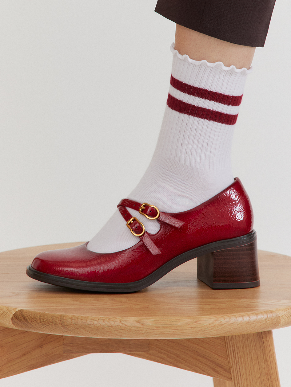 Red patent leather shoe with double buckle on wooden stool, white socks with red stripes and brown trouser hem
