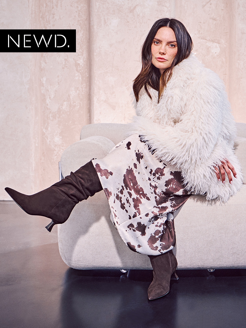 Modelka v bílé kožichové bundě a sukni s motivem kravské kůže sedící na šedé pohovce, má na sobě hnědé semišové boty.