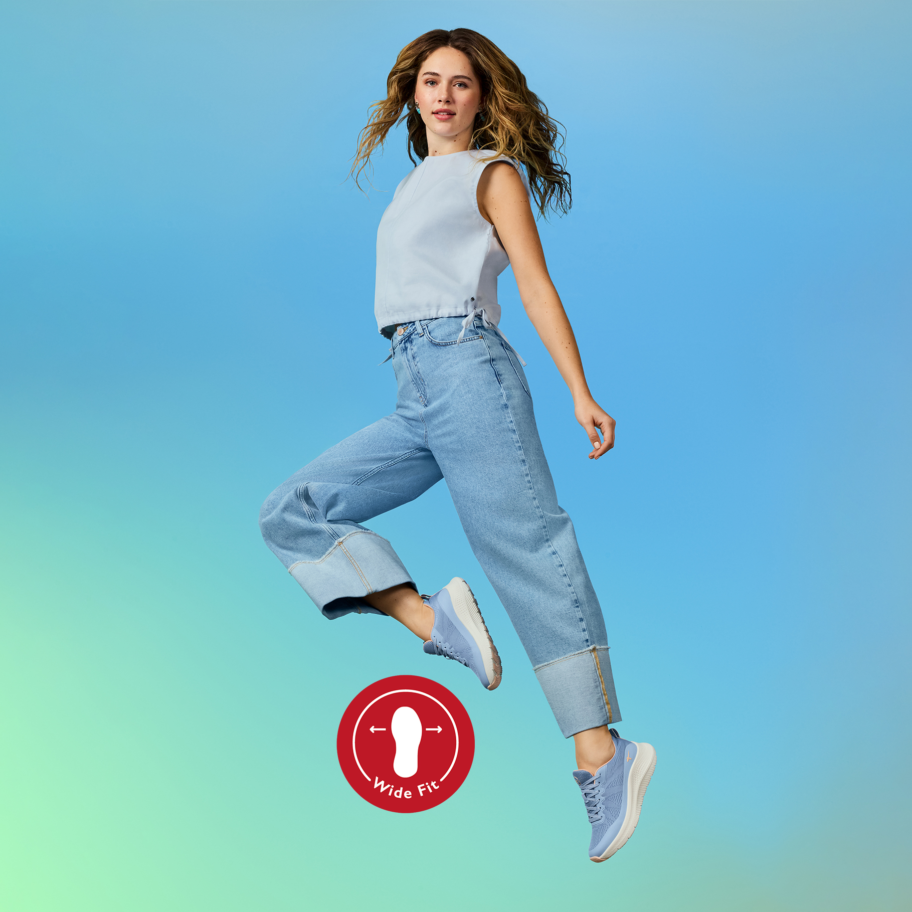 Junge Frau in wei&szlig;em Top und Jeans springt dynamisch vor t&uuml;rkisfarbenem Hintergrund, mit Wide-Fit-Logo unten links.