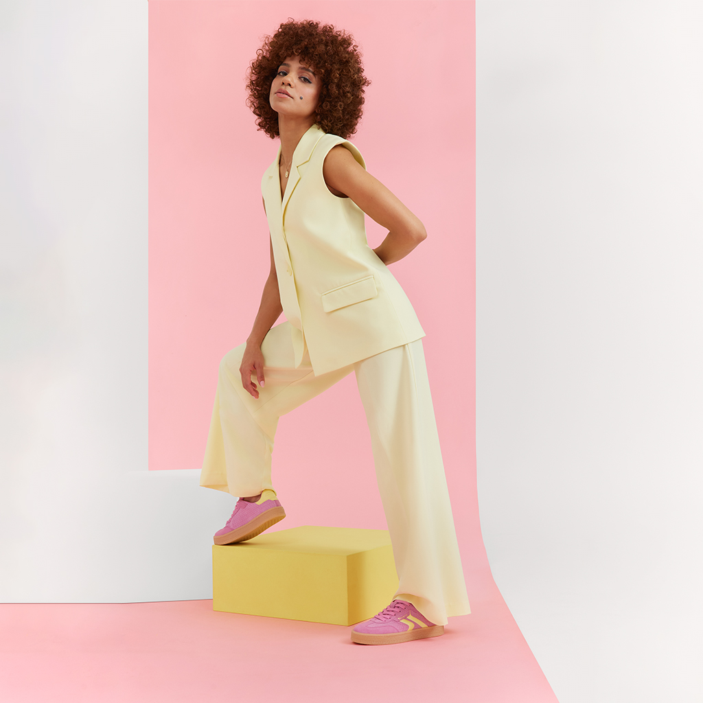 Modell i cremef&auml;rgad byxdress och rosa sneakers poserar dynamiskt mot en rosa bakgrund, med lockigt h&aring;r och en modern look.