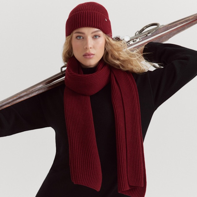 A skier in a black outfit with a wine-red hat and scarf is wearing skis over her shoulder against a white background.