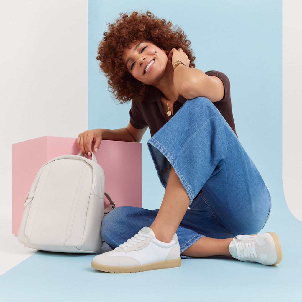 Femme aux cheveux boucl&eacute;s en jean et t-shirt marron posant en souriant &agrave; c&ocirc;t&eacute; d&rsquo;un sac &agrave; dos blanc sur un fond aux couleurs pastel.