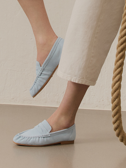 Frauenbeine in beiger Hose tragen hellblaue Loafer aus Leder, neben einem dicken Seil auf hellem Hintergrund.