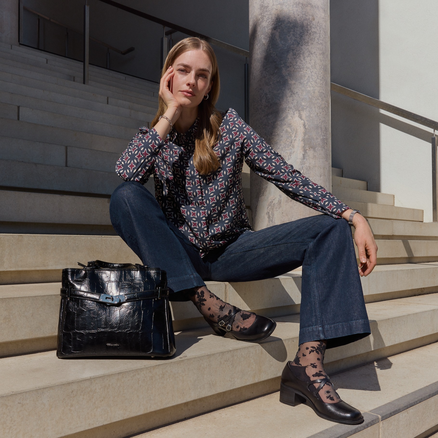 Tenue à la mode avec un chemisier à motifs, un jean et des chaussures noires, assise sur des marches avec un sac à main noir au soleil