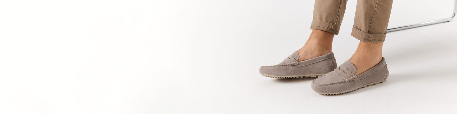 Gr&aring; mockamokasiner burna med beige byxor mot vit bakgrund.