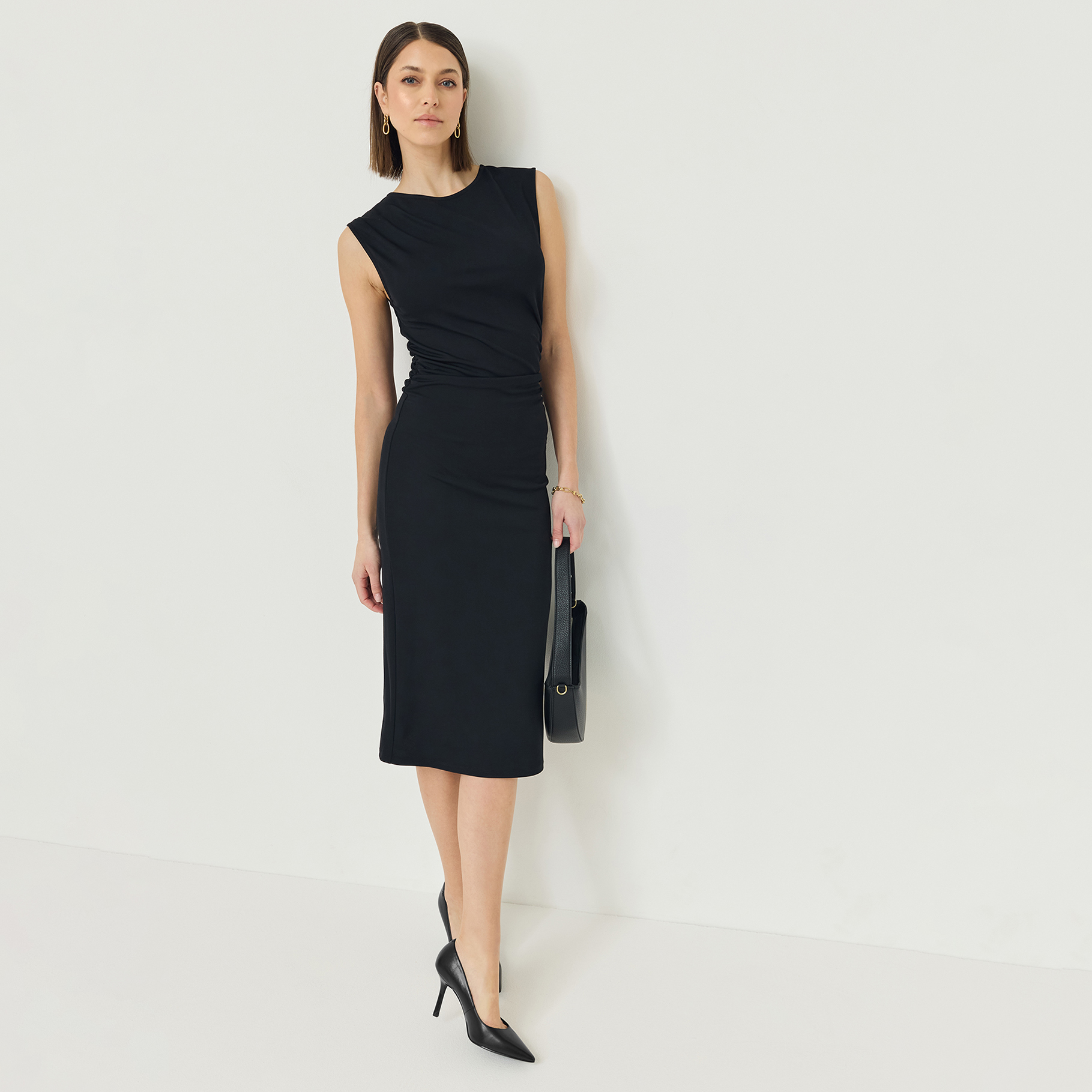 Kvinna i svart &auml;rml&ouml;s midikl&auml;nning med rynkdetalj, svarta pumps och handv&auml;ska, med guldf&auml;rgade &ouml;rh&auml;ngen mot vit bakgrund.