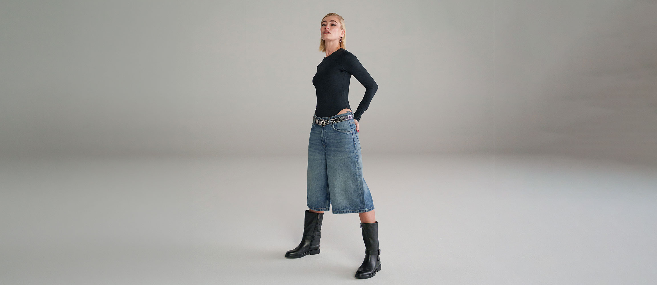 Woman in black long-sleeved shirt, wide denim culottes, belt and black knee-high boots posing against a grey background.